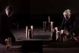 Inszenierungsfoto von Srebrenica, links im Bild Jens Harzer, rechts Vernesa Berbo, die Holzscheite senkrecht wie Grabstelen aufstellen. Beide sind ganz in schwarz gekleidet.