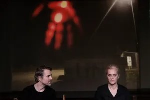 Inszenierungsfoto von Srebrenica, links im Bild Jens Harzer, rechts Vernesa Berbo, die zum Publikum gewandt auf der Bühne sitzen, Harzer blickt zu Berbo, sie schaut nach vorne unten. Sie sind beide in schwarze gekleidet. Im Hintergrund ist eine Projektion eines Fotos zu sehen, darauf eine Laterne und ein Licht, das im unscharfen Zustand aussieht wie eine blutverschmierte Hand.