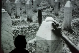 Inszenierungsfoto von Srebrenica, links im Bild die Umrisse von Jens Harzer vor einer Projektion eine Fotos im Hintergrund. Auf dem Foto sind Grabsteine in Form von Stelen zu erkennen sowie eine Hand, die einen dieser berührt. 