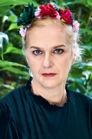 Vernesa Berbo in schwarzem Oberteil mit Blumenschmuck in den Haaren, im Hintergrund sind unscharf Pflanzen zu erkennen. 