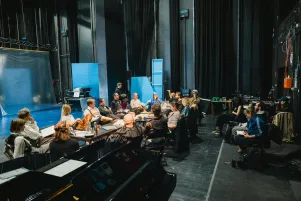 Blick auf den Regietisch, ein Großteil des Teams sitzt daran, im Hintergrund linksseitig sind transparente Stellwände zu erkennen. 