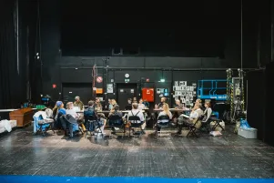 Das Team ist von der Bühne aus fotografiert worden. Sie sitzen lesend und gestikulierend an einem langen Tisch auf der Probebühne. 