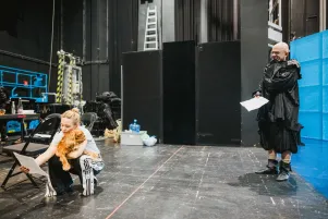 Schnappschuss von Pauline Knof, die in der Hocke sitzend sowohl ein Textblatt, als auch einen kleinen Hund festhält. Mit etwas Abstand schaut Veit Schubert im Probenkostüm schmunzelnd auf sie. 