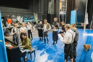 Das Ensemble hat sich um ein Klavier verwandelt, sie halten Notenzettel in der Hand. Der Boden ist leuchtend blau ausgelegt. 