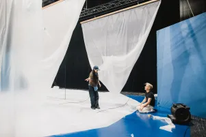 Transparente Folien hängen von der Decke, der Bühnenboden ist zu einem großen Teil Blau ausgelegt, der restliche Teil ist mit der Folie ausgestattet. Auf dem Boden sitzt der Bühnenbildner, der zu seiner Kollegin aufschaut.