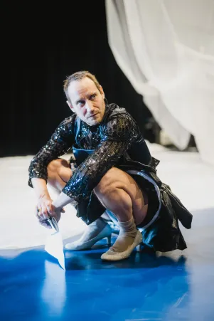 Schnappschuss von Sebastian Zimmler im Probenkostüm. Er hockt auf dem blauen Boden, hält ein Textbuch in der Hand und schaut über seine linke Schulter.