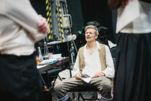 Schnappschuss von Marc-Oliver Schulze. Er sitzt im Probenkostüm aus weißem Hemd, weste und brauner Hose auf einem Stuhl, im vorderen Bildteil sind zwei Körper zu erkennen, zu denen er zu sprechen scheint. 