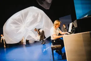 Rechts im Vordergrund spielt eine Person Klavier, während im hinteren Bildausschnitt ein großer Plastikballon aufgeblasen wird. 