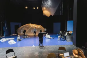 Schnappschuss mit Blick auf die Probebühne. In der Mitte steht der Regisseur, vereinzelt stehen Stühle herum und liegen Folien auf dem Boden. Es ist ein beleuchteter Folienballon zu erkennen. 