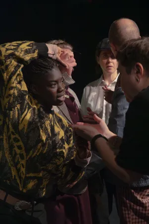 Joyce Sanhá und Stas Zhrykov im Gespräch. Sie hat ihren rechten Arm über ihren Kopf gelegt, im Hintergrund sind weitere Ensemblemitglieder zu erkennen. 