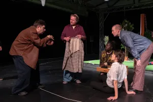 Der Regisseur spricht zum Ensemble, er scheint mit seiner Gestik eine Figur nachzumachen. 