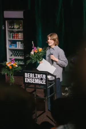 Amelie Willberg vor einem Pult des Freundeskreises im Bühnenbild von "Die Verstreuten". In der linken Hand hält sie ein Mikrofon, in der Rechten einen großen Blumenstrauß und einen weißen Umschlag. 