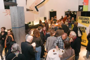 Ein gut gefülltes Foyer, Menschen stehen herum, unterhalten sich, trinken. Zwischendrin sind die Kunstobjekte von Alex Wissel zu erkennen. 