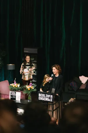 Tena Štivičić steht vor einem hohen Tisch mit dem Logo des Freundeskreises im Bühnenbild von "Die Verstreuten". Sie spricht in ein Mikrofon, neben ihr sind ihre Tochter und ein Vorstandsmitglied des Freundeskreises zu erkennen. 