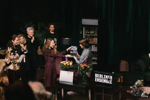 Amelie Willberg erhält den Nachwuchsförderpreis von Kathleen Morgeneyer. Sie stehen im Bühnenbild von "Die Verstreuten", um sie herum applaudieren die Jurymitglieder und Intendant Oliver Reese. 