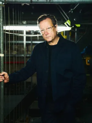 Portrait von Matthias Brandt in einer der Galerien zwischen Seilzügen. Er trägt ein dunkles Outfit und hält sich mit der rechten Hand an einer Metallstange fest.