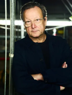 Portrait von Matthias Brandt in naher Position, hinter ihm ist eine Halogenleuchte zu erkennen, außerdem einige Seilzüge. Er verschränkt die Arme vor der Brust. 