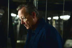 Portrait von Matthias Brandt im Profil. Im Hintergrund sind Leuchten und einzelne Seilzüge zu erkennen. Er trägt eine Brille und schaut leicht nach unten. 