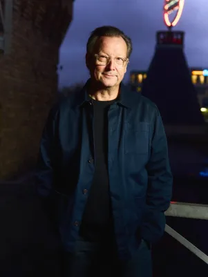 Portrait von Matthias Brandt auf dem Dach des Berliner Ensembles. Es ist dunkel, im Hintergrund leuchtet das Logo auf dem Turm. Er schaut direkt in die Kamera.