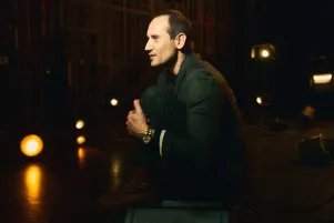 Portrait von Sebastian Zimmler auf der Bühne. Er ist im Profil zu erkennen, sitzt auf einem Scheinwerfer, hat die Knie angezogen und die Arme um sie gelegt. 