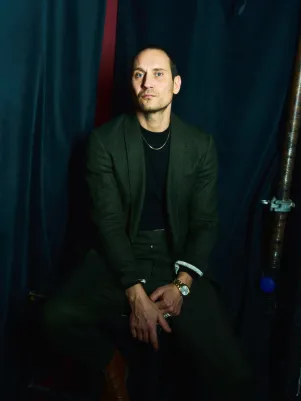 Portrait von Sebastian Zimmler. Er sitzt, trägt einen dunklen Anzug, eine Kette blitzt an seinem Kragen hervor. Hinter ihm ein dunkler Vorhang. 