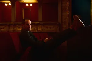 Foto von Sebastian Zimmler im Zuschauerraum des Großen Hauses. Im Hintergrund sind die Parkettlogen zu erkennen, das Bild ist rötlich gefärbt. Sebastian Zimmler sitzt auf einem Samtstuhl, legt die Füße auf den Vordersitz und schaut in die Kamera.