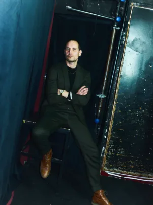 Sebastian Zimmler hockt auf einem Schemel, hat die Arme vor der Brust verschränkt und schaut direkt in die Kamera. 