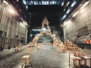 Blick auf die große Bühne: Links und rechts stehen hölzerne Stühle, im hinteren Teil ist ein Rundbogen aus Holzstühlen gebaut worden. 