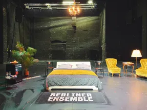 Auf der Bühne des großen Hauses steht ein Doppelbett davor ein Bett mit dem Aufdruck "Berliner Ensemble", außerdem eine Kunstpalme und ein Nachttisch mit Lampe. Rechts stehen zwei gelbe Samtsessel samt kleinem Beistelltisch. 