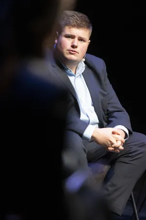 Schnappschuss von Johannes Volkmann. Er trägt einen dunklen Zweiteiler samt weißem Hemd, hat die Hände ineinander verschränkt und scheint aufmerksam zuzuhören. Seine Miene wirkt verschlossen.  