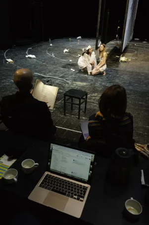 Blick aus dem Off auf die Bühne: Es ist ein aufgeklappter Laptop samt Textbuch zu erkennen, davor  die Soufflage und auf der Bühne zwei Spieler:innen in knienden Posen, sie tragen Probenkostüme.