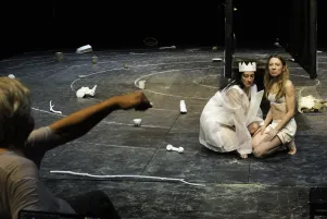 Constanze Becker und Kathleen Morgeneyer knien voreinander auf der Bühne, sie tragen weiße Probenkostüme, um sie herum sind weiße Objekte zu erkennen. Im linken Bildrand regt Johan Simons gestikulierend die Faust.