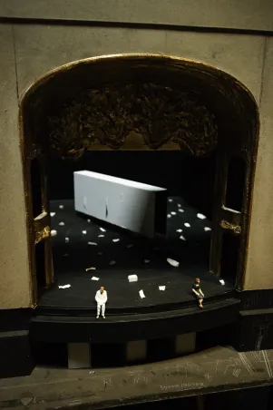 Blick aus dem zweiten Rang auf die Bühne des Großen Hauses. Darauf sind ein weißer Container und viele weiße  Objekte zu erkennen, an der Rampe stehen und sitzen zwei Ensemblemitglieder.