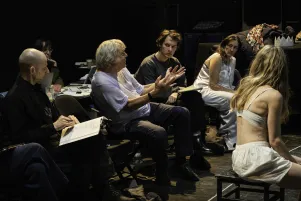 Johan Simon scheint mit dem Regieassistenten zu diskutieren. Um sie herum sitzen einige Teammitglieder. 