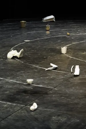 Es sind weiße Objekte zu erkennen, die auf der Bühne verteilt liegen. Darunter eine halbe Büste, ein Telefonhörer, ein Gipsfuß. 