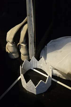 Schnappschuss: Es sind auf dem Boden liegende Requisiten zu erkennen, darunter weiße Stiefel, eine weiße Krone und eine Art Deckel.