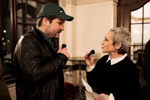 Max Czollek und Marion Brasch nehmen im Foyer des Großen Hauses die Begrüßung auf. Sie sind im Profil zu erkennen, einander zugewandt und halten kleine Mikrofone in den Händen. 