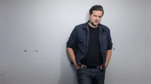 Christian Fuchs steht an eine weiße Wand gelehnt, hat seine Hände in den Hosentaschen. Er trägt Jeans, schwarzes Shirt und eine blaube Jacke darüber. Er schaut in die Kamera.