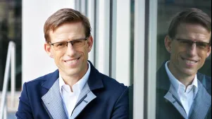Fritz Breithaupt in blauem Jackett. Er lehnt an einer verspiegelten Wand, der Hintergrund ist unscharf. Er lächelt, schaut in die Kamera und trägt Brille. 