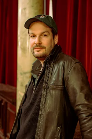 Portrait von Max Czollek im Profil. Er steht vor einem roten Samtvorhang und  schaut ernst in die Kamera, die Arme hat er hinter sich abgestützt. 