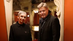 Robert Habeck und Marion Brasch stehen in einer Loge, im Hintergrund ist der Zuschauerraum zu erkennen. Sie tragen beide dunkle Kleidung und schauen direkt in die Kamera.