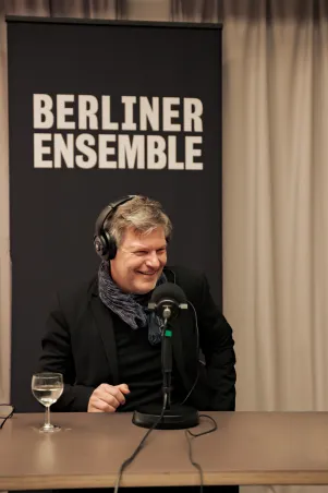 Robert Habeck sitzt im Nebenraum der Kantine am Mikrofon. Er ist seiner Gesprächspartnerin Constanze Becker, die nicht im Bild ist, zugewandt und lacht. 