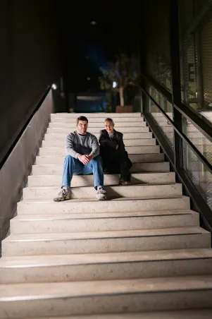 Simon Dömer und Marion Brasch sitzen auf der Betontreppe des Neuen Hauses und schauen direkt in die Kamera. 