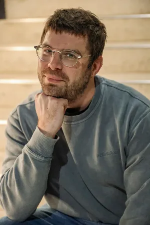 Portrait von Simon Dömer auf der Treppe sitzend. Er lehnt das Kinn auf seine Hand auf. 
