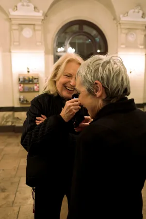 Maren Kroymann und Marion Brasch unterhalten sich im Foyer des Großen Hauses. Sie halten Mikrofone in der Hand und lachen. Marion Brasch ist der Kamera mit dem Rücken zugewandt, es ist ihr Profil zu erkennen. 