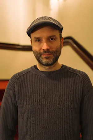 Portrait von Marc-Uwe Kling im Treppenhaus zum ersten Rang. Er trägt eine Cap und einen braunen Pullover, das Foto ist leicht von unten aufgenommen, er schaut direkt in die Kamera. 