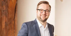 Oliver Nachtwey in weißem Hemd und blauem Sacko. Er trägt Brille und lacht in die Kamera. Im Hintergrund ist eine weiße Wand und an der linken Seite unscharf eine Holzvertäfelung zu erkennen.