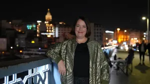 Phoebe Gaa auf einer Brücke bei Nacht, im Hintergrund eine beleuchtete Skyline einer Stadt. Sie trägt einen dunkle Oberteil, darüber eine goldene Jacke. Sie hat einen dunkelbraunen Bob.