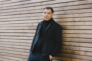 Max Hopp lehnt an eine Holzwand gelehnt, die Hände in den Hosentaschen.