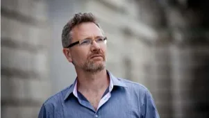 Mann mit Brille und hellblauem Hemd blickt mit dem Kopf leicht geneigt nach vorne oben. Der Hintergrund ist unscharf.
